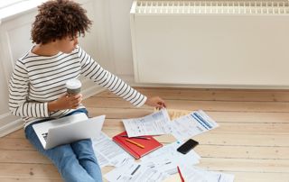 Une femme assise au sol près du radiateur avec son ordinateur et des papiers